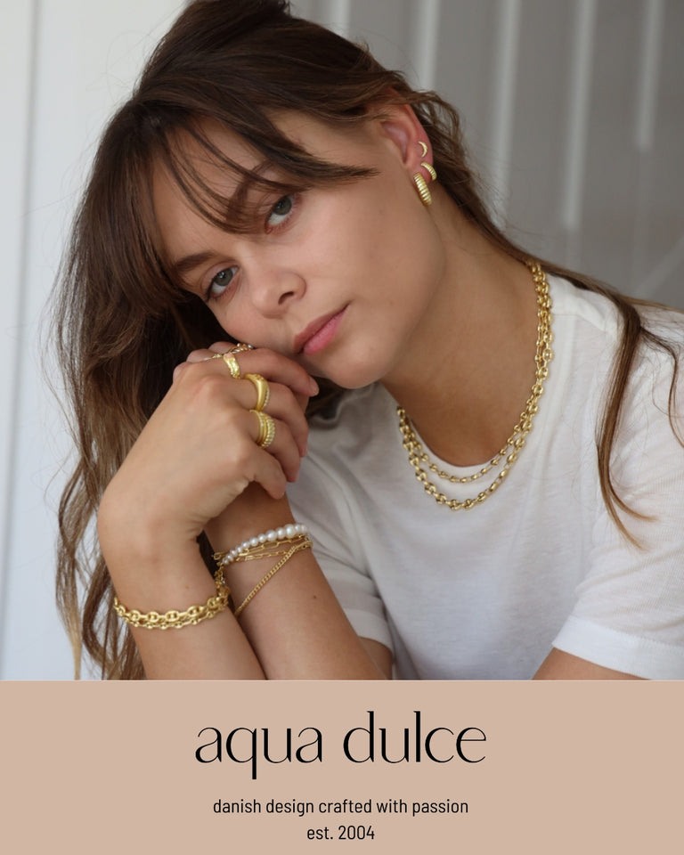 Aqua Dulce smykkemodel iført guldøreringe, armbånd, halskæder og ringe, forsidebillede