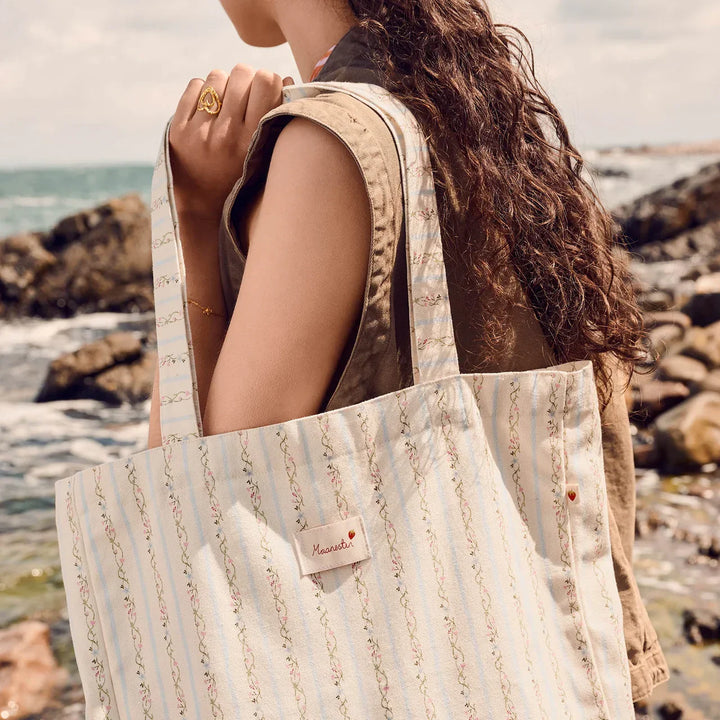 maanesten-canvas-totebag-summer-stripes-blue worn outdoors at the coast, light blue stripes and embroidered details. Model 4121-one size