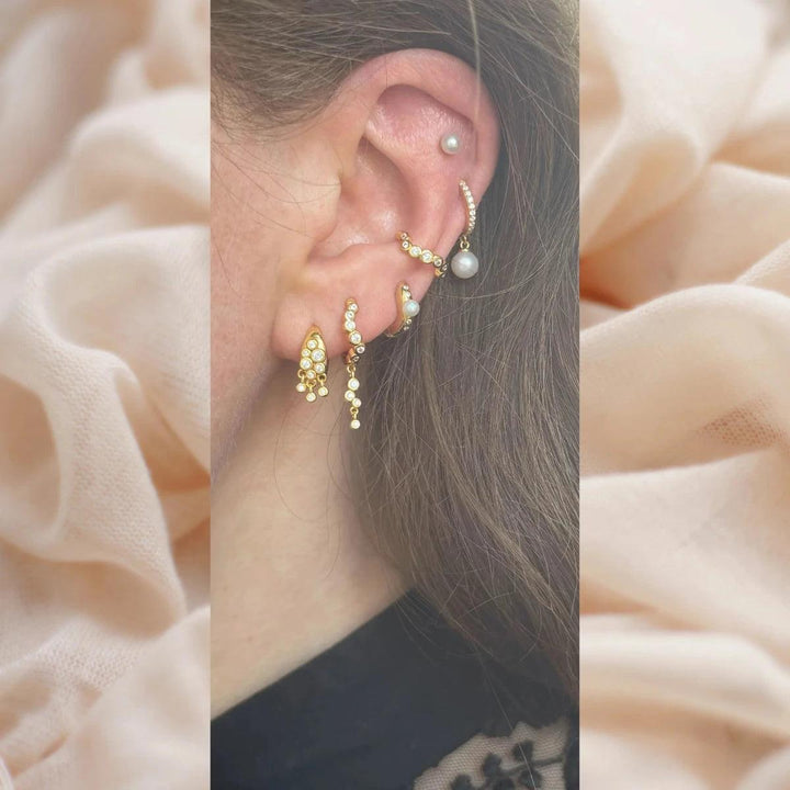 Sirona Huggie shown on ear with multiple gold earrings and pearl details, in light surroundings. Model fw2405-2