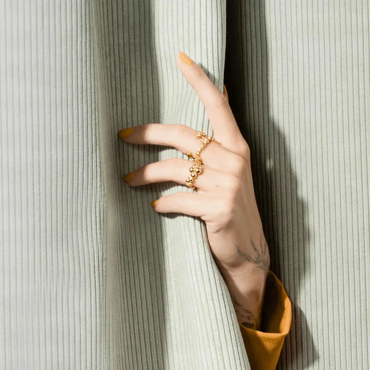 Vincent Orb Inferior Gilt Ring, shown on hand with yellow nail polish in front of pale green velvet curtain. Model VJ215-URG-001-2