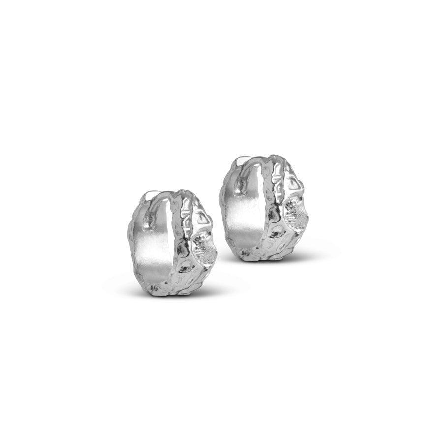 Enamel Nela Hoops shows a pair of small, silver-colored earrings with an organic, raw surface. Model E240SM-E240SM-1